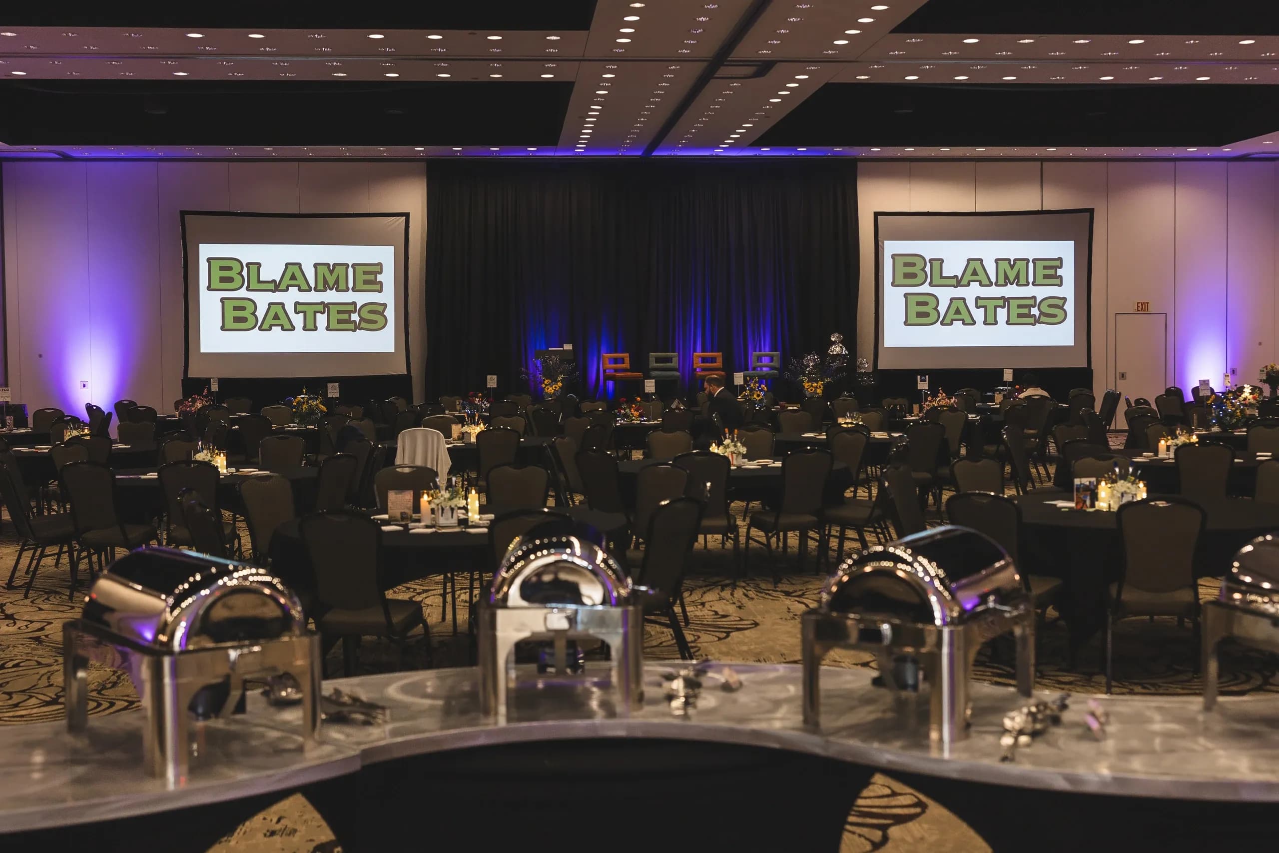 The gala ballroom set for dinner, with Blame Bates branding on two screens.