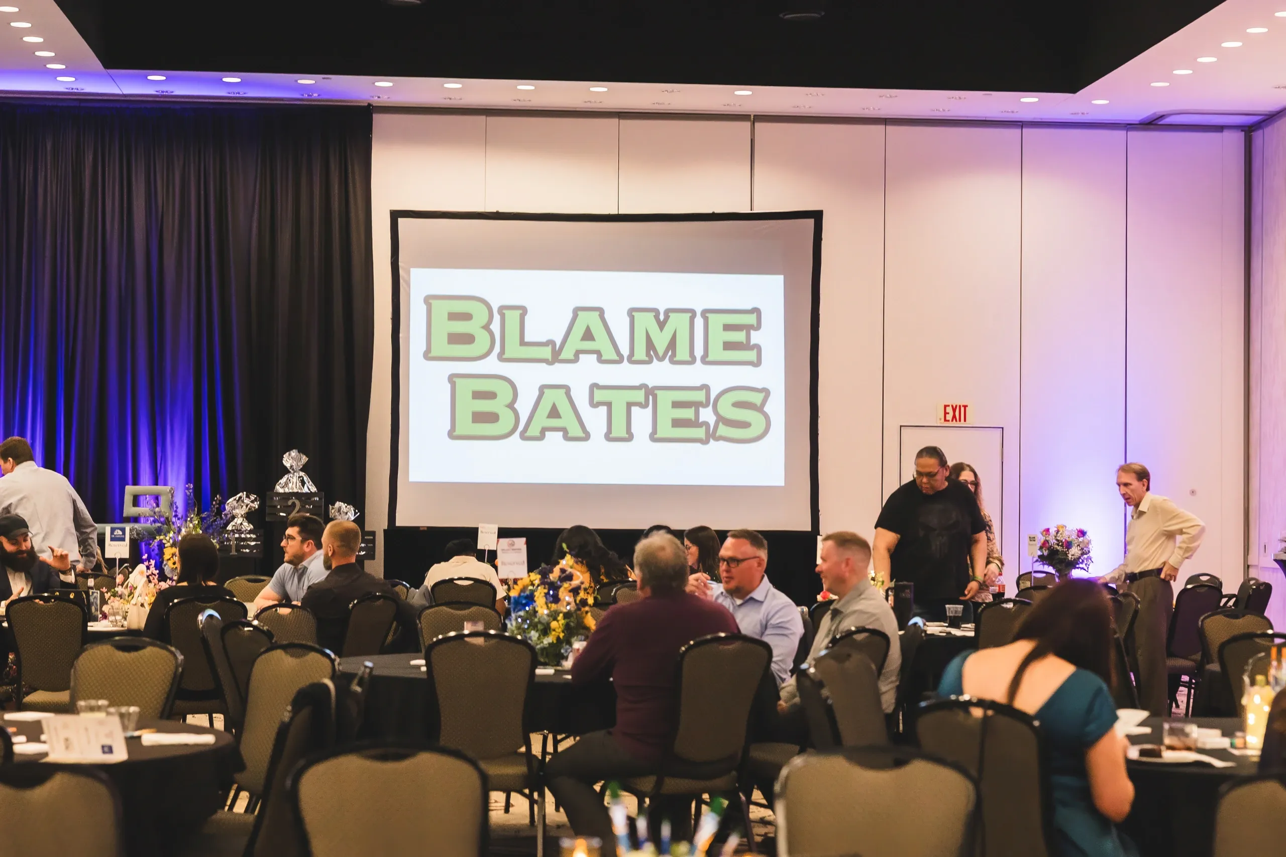 Guests seated at round tables during the Paint the Night Away gala, with the Blame Bates logo projected on screen.